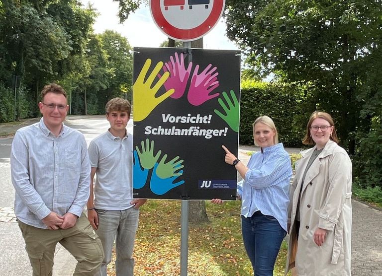 Vier Personen vor Schulweg-Schild mit Handabdrucken und Warnsymbolen für Fußgänger und Autos.