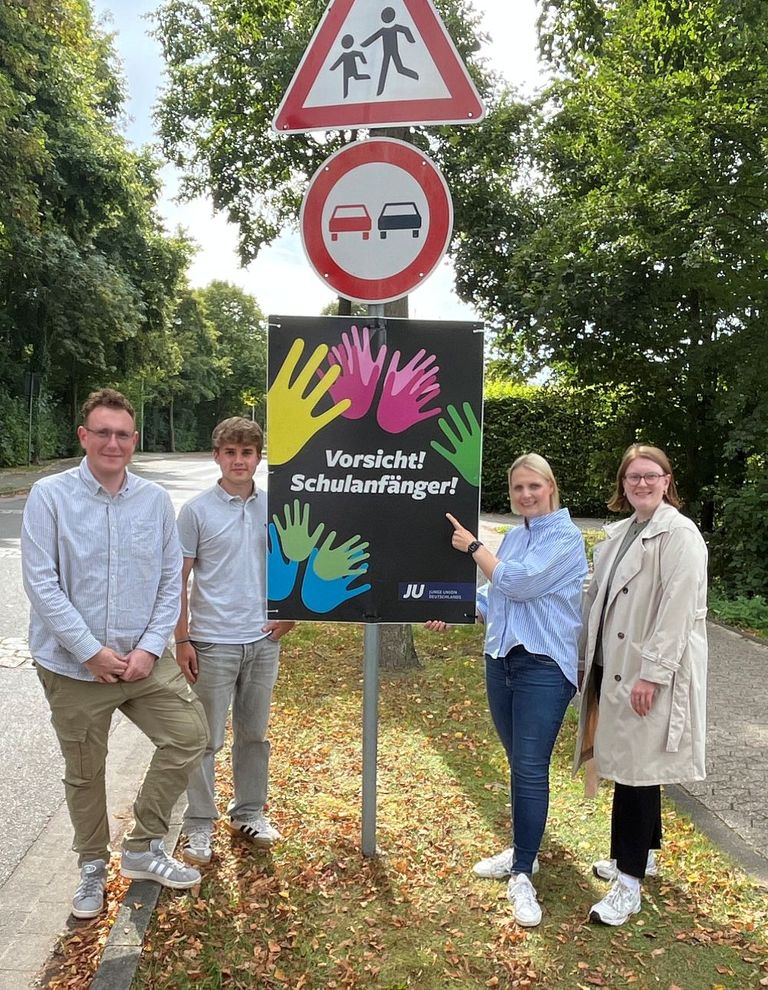 Vier Personen vor Schulweg-Schild mit Handabdrucken und Warnsymbolen für Fußgänger und Autos.